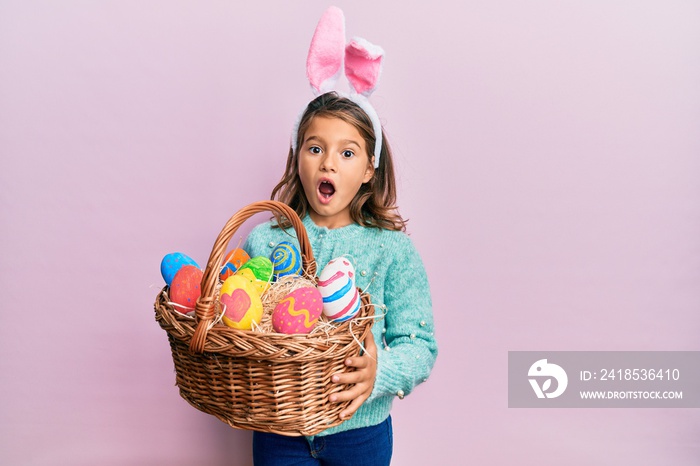 Little beautiful girl wearing cute easter bunny ears holding wicker basket with colored eggs celebra