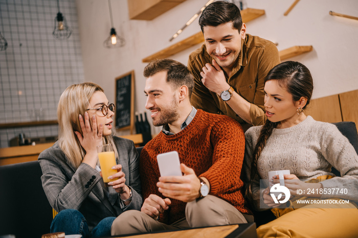 surprised woman looking at man holding smartphone near friends