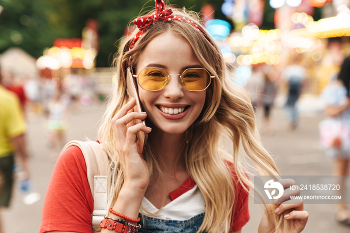 Smiling pleased young cheery blonde woman in amusement park talking by mobile phone.