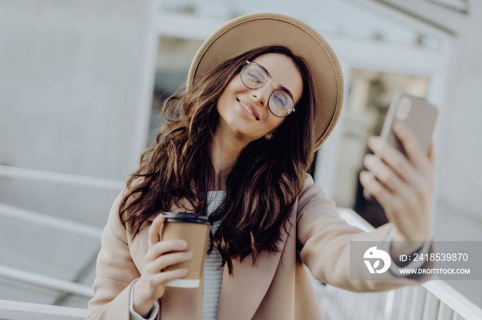 Beautiful young woman wearing a coat taking a selfie with mobile phone, holding takeaway coffee cup 