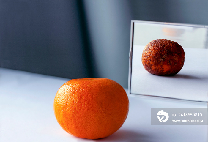 А fresh orange on the table is reflected in the mirror as rotten фтв spoiled fruit. Conceptual photo