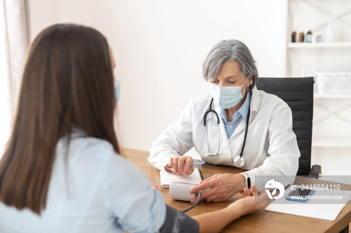 Senior mature female doctor wearing a lab coat and a face mask, using a blood pressure machine or sp