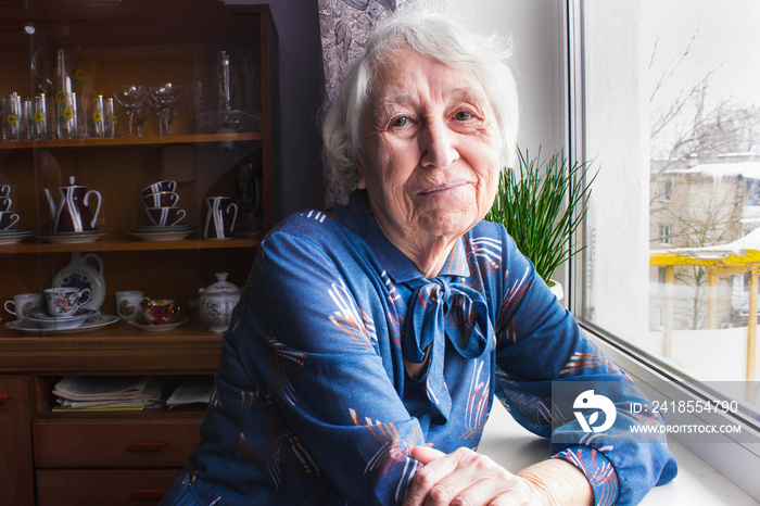 Old lonely woman sitting near the window in his house.