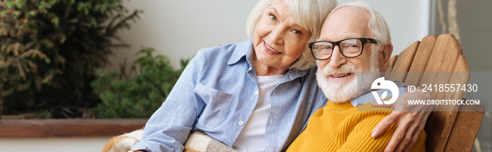 smiling senior wife hugging husband and looking at camera on terrace on blurred background, banner