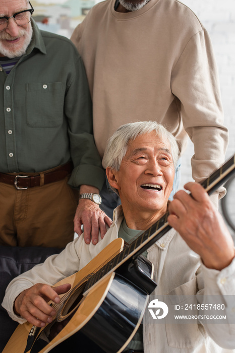 一位年长的亚洲男子在家里的异族朋友附近弹原声吉他。