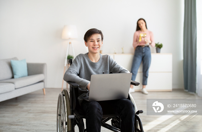 Happy handicapped teen boy in wheelchair with laptop computer indoors, his mother standing on backgr