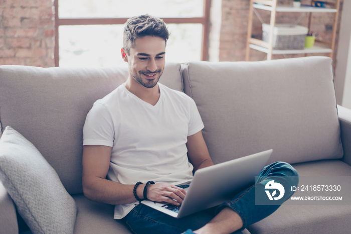 一个年轻迷人、面带微笑的家伙正在浏览他的笔记本电脑，坐在家里舒适的米色沙发上。