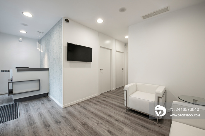 Waiting room in a clinic with tv, white walls and sofas and grayish wooden floors