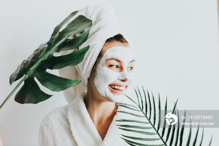 Young beautiful woman in face mask of therapeutic white mud and tropical leaves. Spa treatment, self