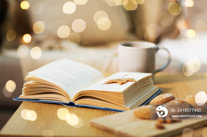 hygge and cozy home concept - book with autumn leaf, cup of tea and oatmeal cookies on wooden table