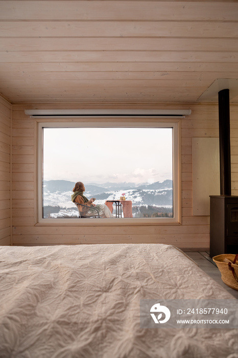 Woman sitting with a drink on terrace of tiny house in the mountains, view through the window from t