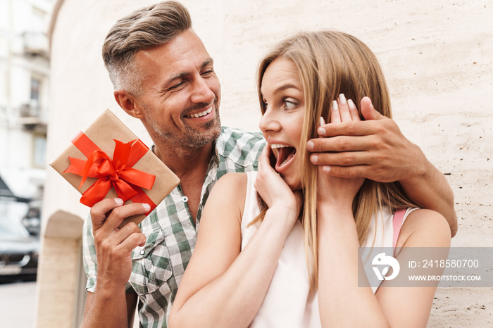 Image of astonished excited couple smiling and holding present box together while standing against w