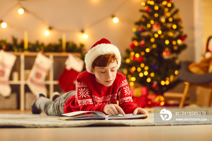 Concentrated boy in red Christmas clothing and hat lying on floor and reading book