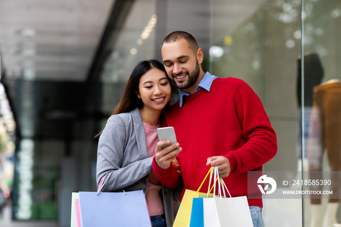 快乐的跨种族年轻夫妇带着礼品袋和手机在shoppin附近在线订购商品