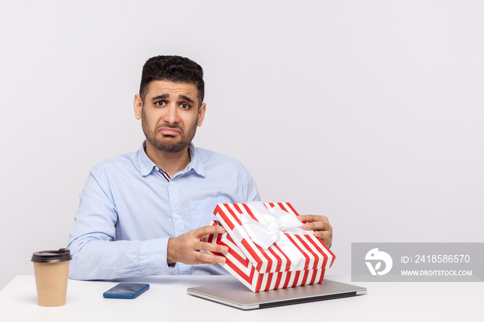 Upset man employee sitting in office workplace, opening gift box and looking at camera with disappoi