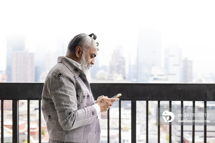 gray hair bearded man looking at smartphone on balcony with city view