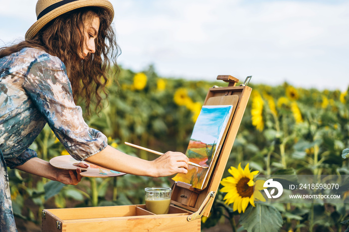 A young woman with curly hair and wearing a hat is painting in nature. A woman stands in a sunflower