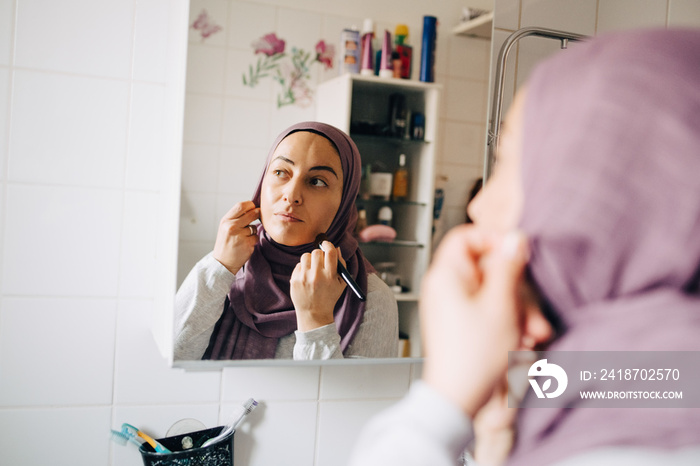 Mature woman in hijab doing make-up while reflecting in mirror at home