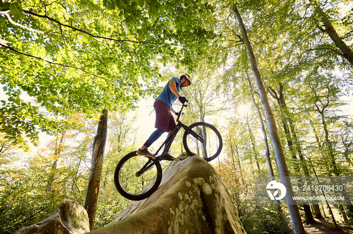 Male professional cyclist balancing on trial bicycle, making acrobatic trick on big boulder in the f