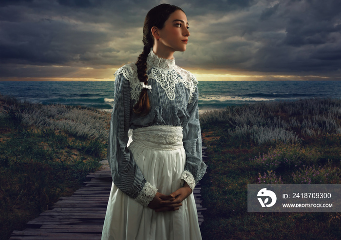 Young victorian girl in white dress and blue striped blouse on the coast at sunset.