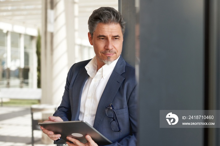 Confident elegant older businessman in suit standing and working with business tablet computer in of