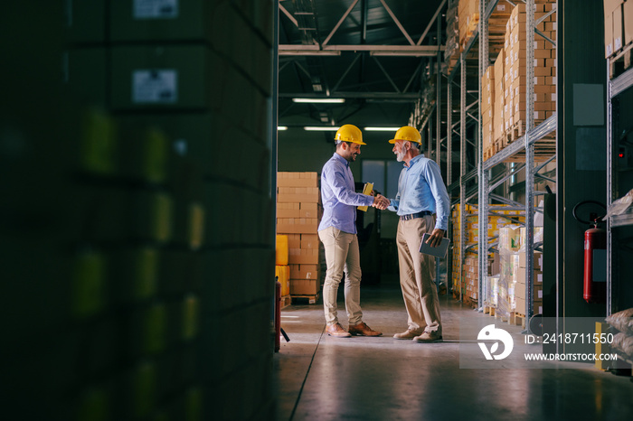 Two businessman with helmets on heads shaking hands for good finished job. Storage interior.