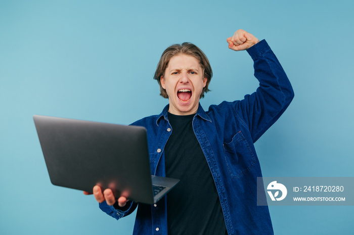 Young man with a laptop in his hands emotionally rejoices in victory with his hand raised on a blue 