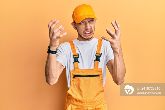 Hispanic young man wearing handyman uniform shouting frustrated with rage, hands trying to strangle,