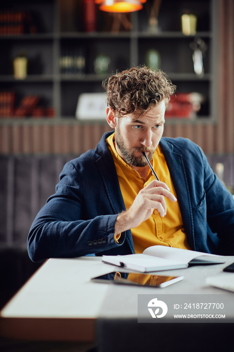 Young serious bearded Caucasian blogger dressed smart casual writing notes in agenda and looking at 