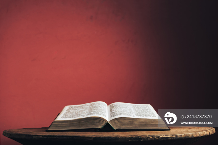 Open Holy Bible on a old brown round wooden table. Beautiful red wall background.