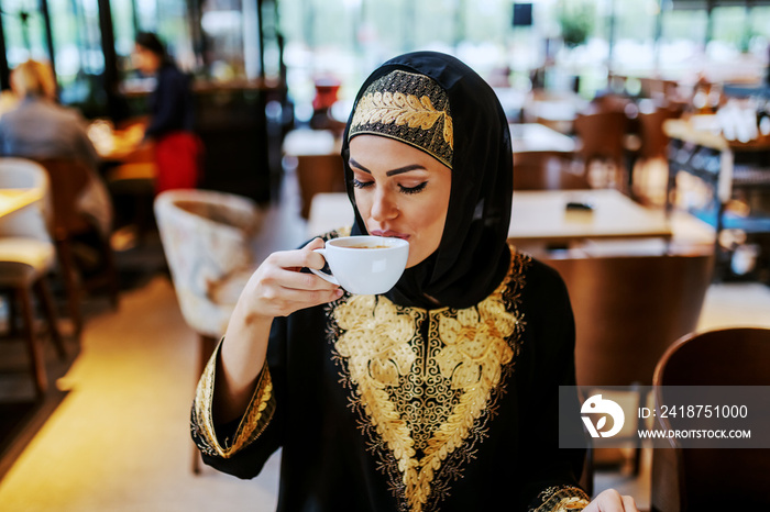 Lovely positive attractive arab woman dressed in traditional wear sitting in cafe alone and enjoying