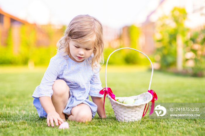 一个穿着蓝色连衣裙的可爱小女孩坐在草坪上，在柳条篮子里收集彩蛋。Eas