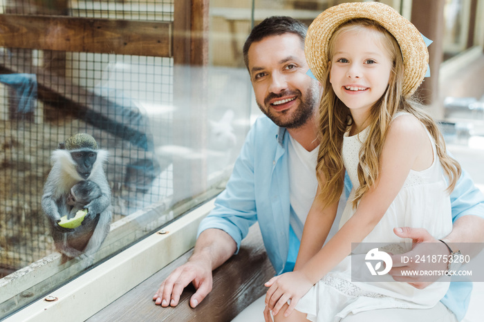happy father and daughter looking at camera near monkey while sitting near window in zoo
