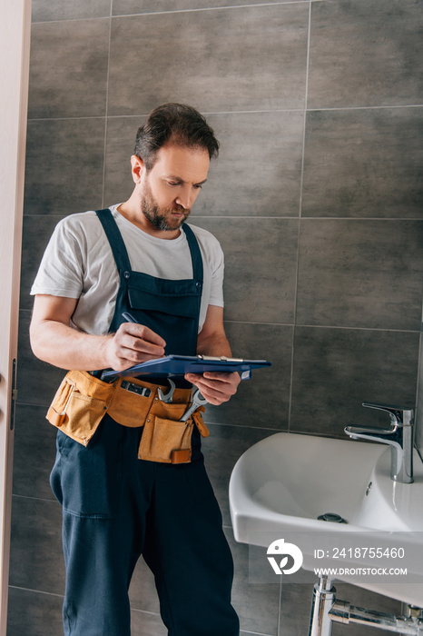 handsome male plumber with toolbelt writing in clipboard near broken sink in bathroom