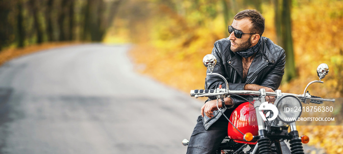 Bearded brutal man in sunglasses and leather jacket sitting on a motorcycle on the road in the fores