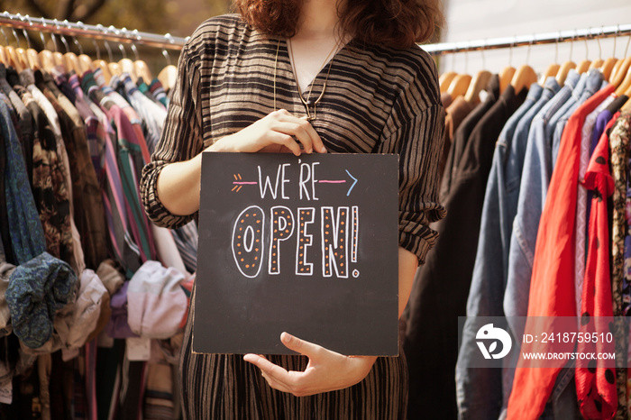 Mid section or woman holding open sign with clothes racks on background