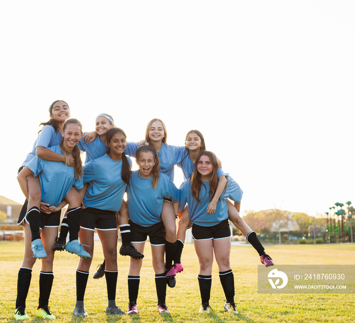 Low angle portrait of girls piggybacking friends while standing on soccer field against clear sky du