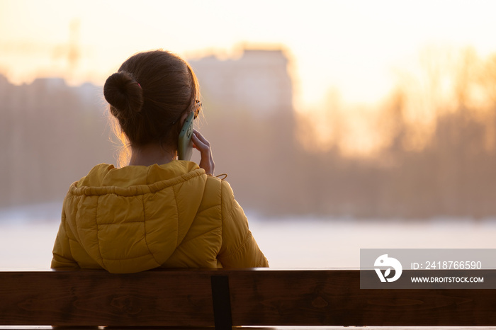 Young pretty woman sitting on a bench talking on her mobile phone outdoors in the evening.