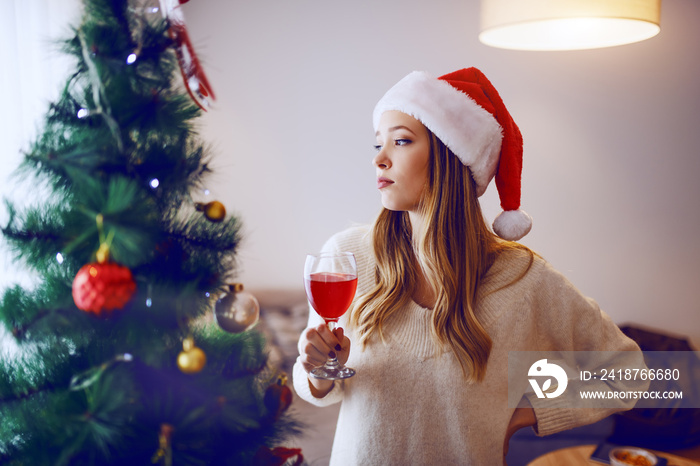 Sad caucasian blonde woman with santa hat on head drinking wine alone and looking trough window. In 