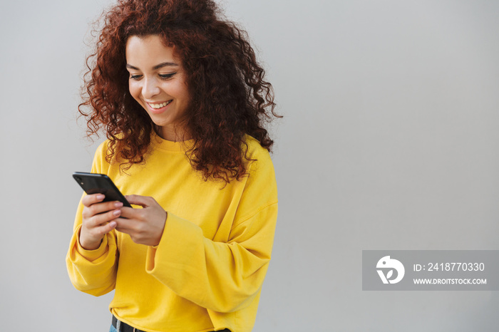 Happy smiling cheerful beautiful curly girl isolated over gray background using mobile phone.