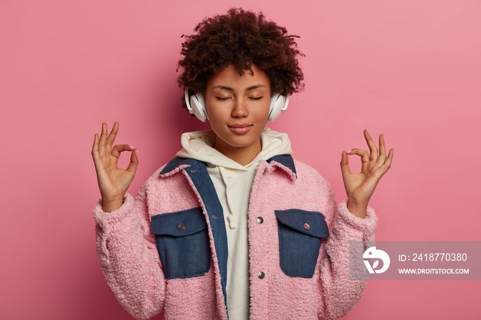 Portrait of dreamy relaxed curly haired woman meditates while listens calm music, makes om gesture, 