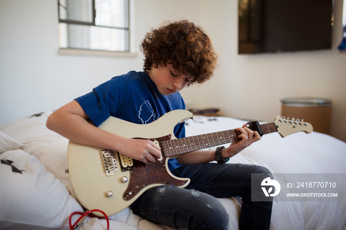 Boy with curly hair playing guitar while sitting on bed at home