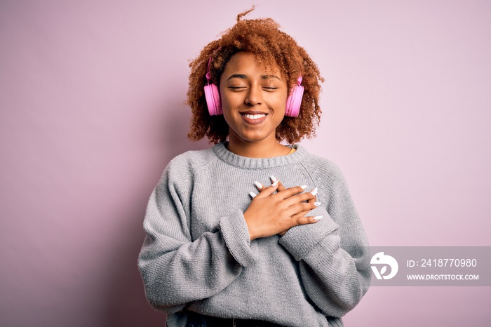 Young African American afro woman with curly hair listening to music using pink headphones smiling w