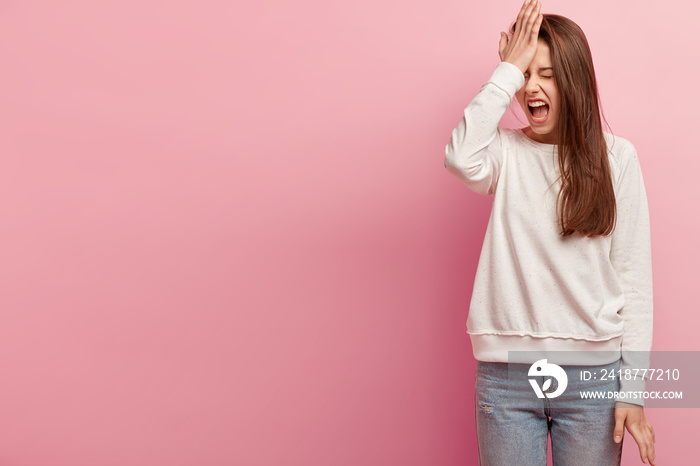 Studio shot of dissatisfied woman slaps hand on forehead, regrets doing bad mistake, screams from de