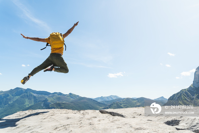 Young Man Jumping on Top of a Mountain Wearing Yellow Backpack.