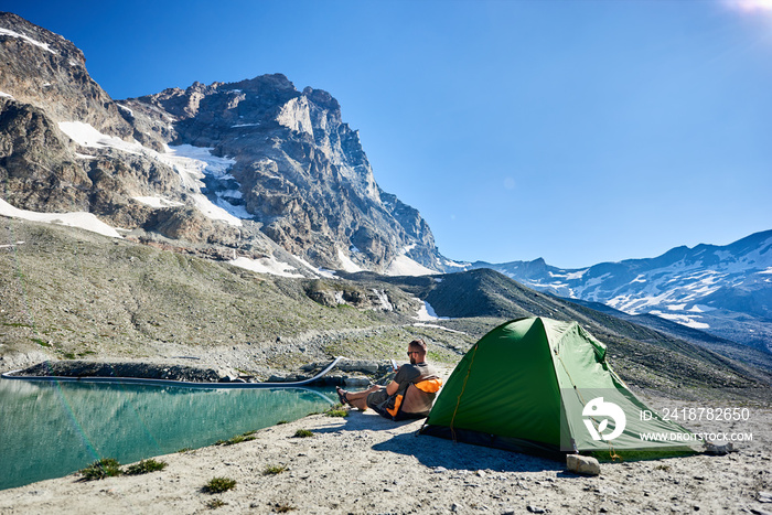 在湖边空旷的岩石高山上，一名男子坐在帐篷附近的椅子上，使用smar的后视图
