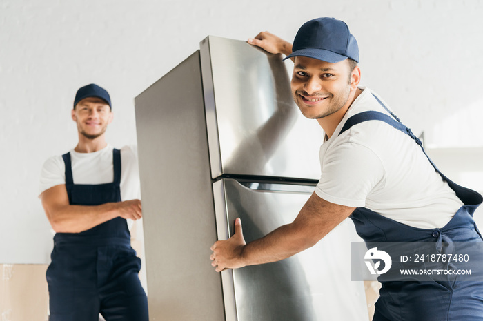 cheerful indian mover in uniform moving fridge near coworker on blurred background