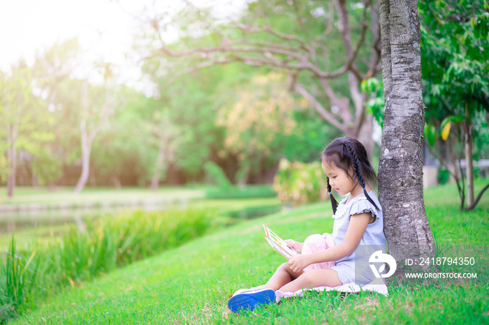 Cute little girl reading a book with a doll in the park