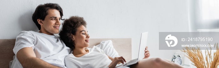 happy african american freelancer using laptop near boyfriend in bed, banner