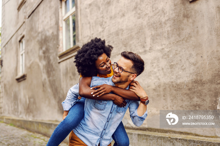 Young smiling overjoyed multicultural hipster couple having piggyback outdoors.